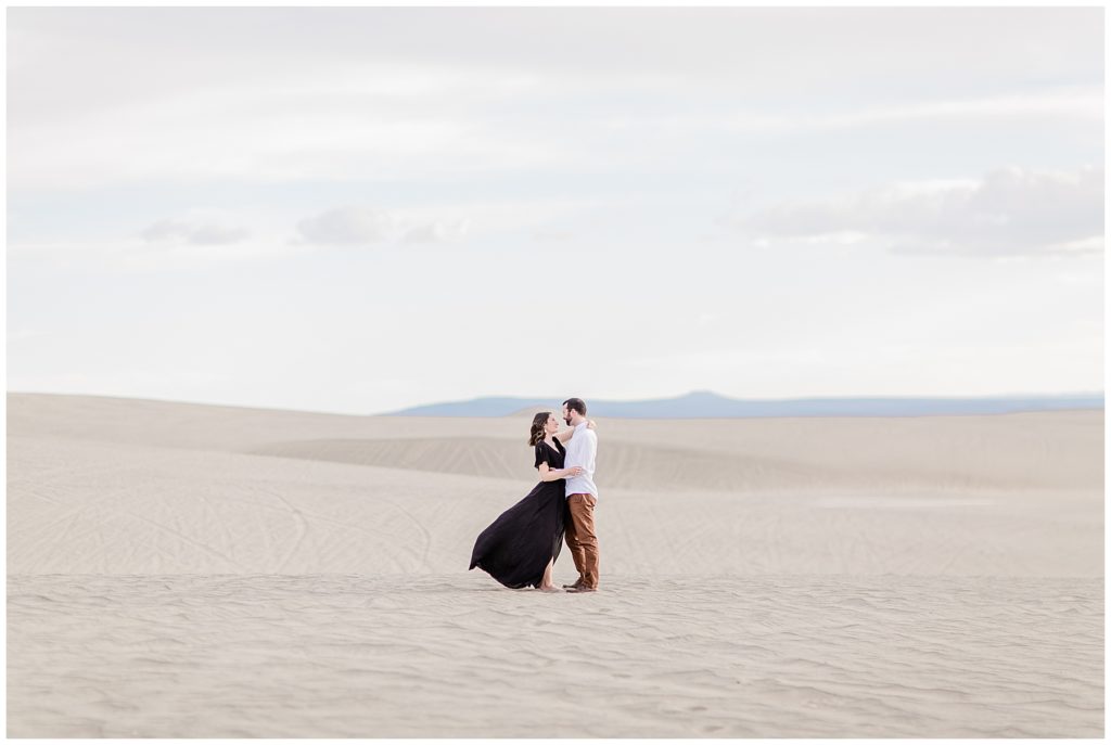 Couple poses in the desert for engagement portraits in Bend, Oregon. She is wearing black flowy dress. He is in a white button down shirt. 